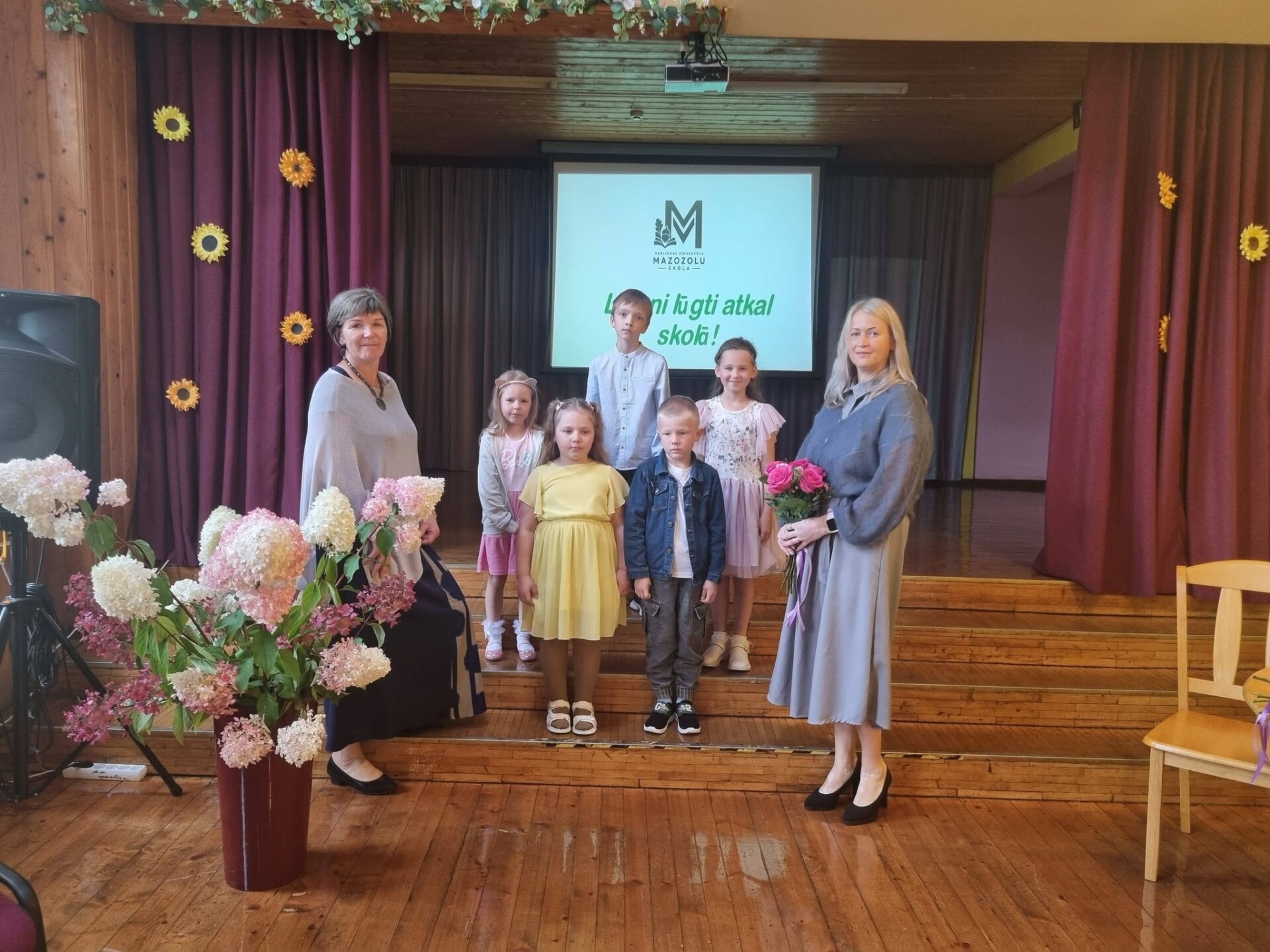 Svinīgajā Zinību dienas pasākumā ir nostājušies sākumsskolas skolēni un divas viņu skolotājas.