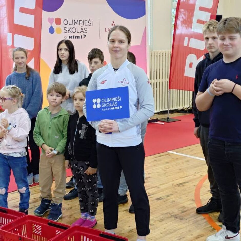 Olimpiete ar projekta plāksni kopā ar skolēniem sporta zālē