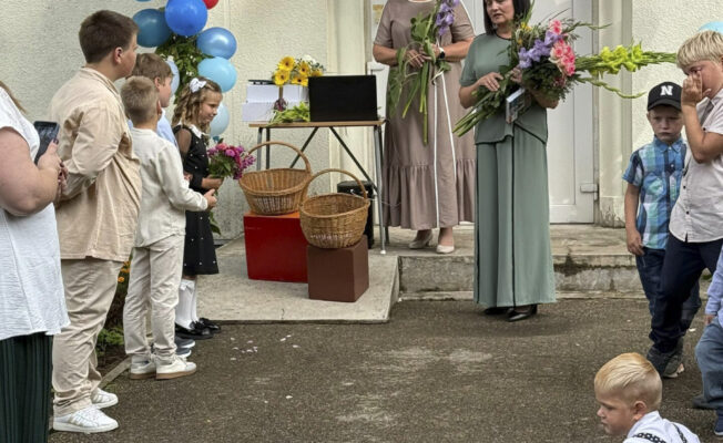 Pie durvīm, kas rotātas ar ballonu arku stāv divas skolotājas, bet priekšpusē ir bērni, kuri stāv un klausās un divi mazuļi, kuri notupušīes spēlējas