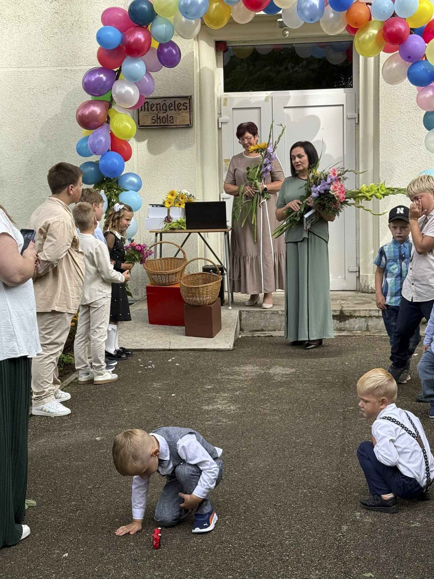 Pie durvīm, kas rotātas ar ballonu arku stāv divas skolotājas, bet priekšpusē ir bērni, kuri stāv un klausās un divi mazuļi, kuri notupušīes spēlējas
